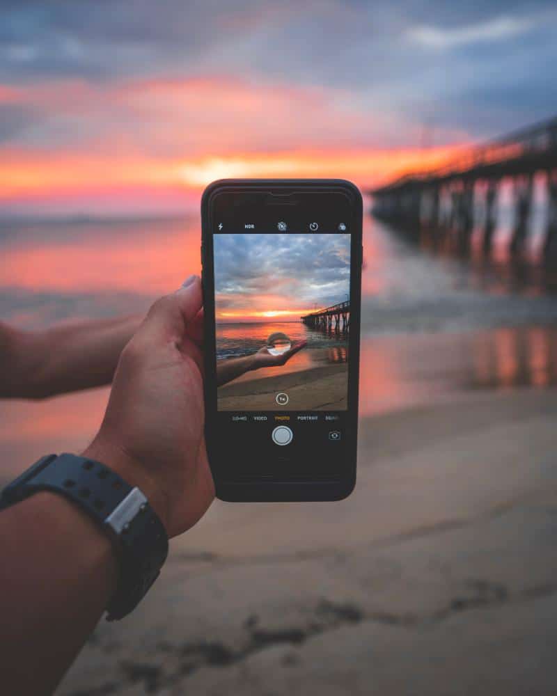 person_holding_black_smartphone_near_seashore_and_dock_under_cloudy_sky_during_golden_hour-scopio